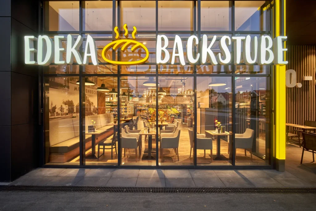 Beleuchtete Glasfassade mit EDEKA-Backstube-Schriftzug bei Dämmerung