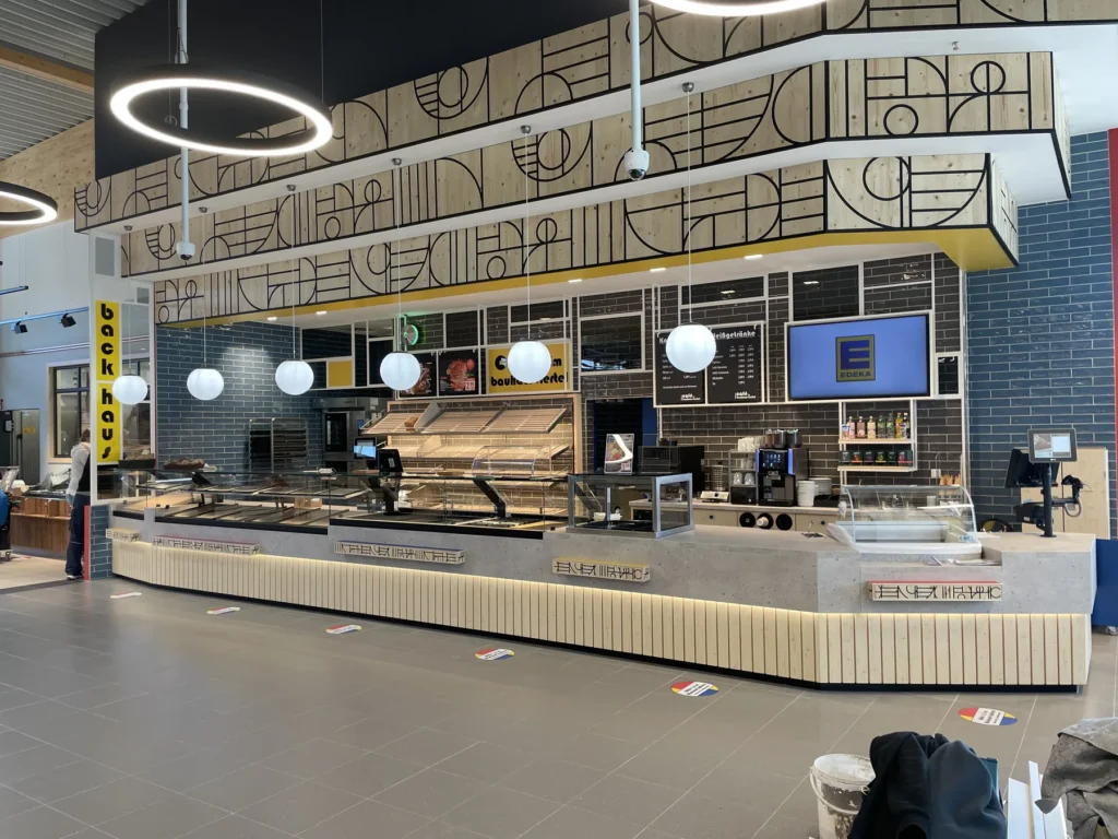 Bedientheke mit Brotregal unter geometrischer Bauhaus-Wandgrafik, EDEKA-Bildschirm im Hintergrund